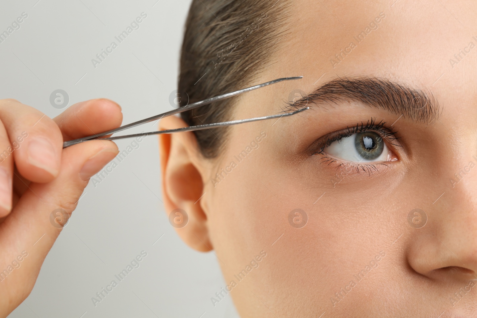 Photo of Eyebrow correction. Young woman with tweezers on light grey background, closeup