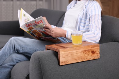 Photo of Glass of juice on wooden sofa armrest table. Woman reading magazine at home, closeup
