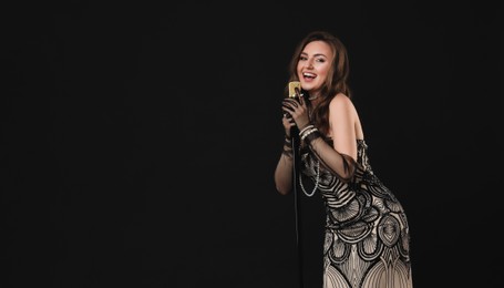 Beautiful young woman in stylish dress with microphone singing on black background, space for text