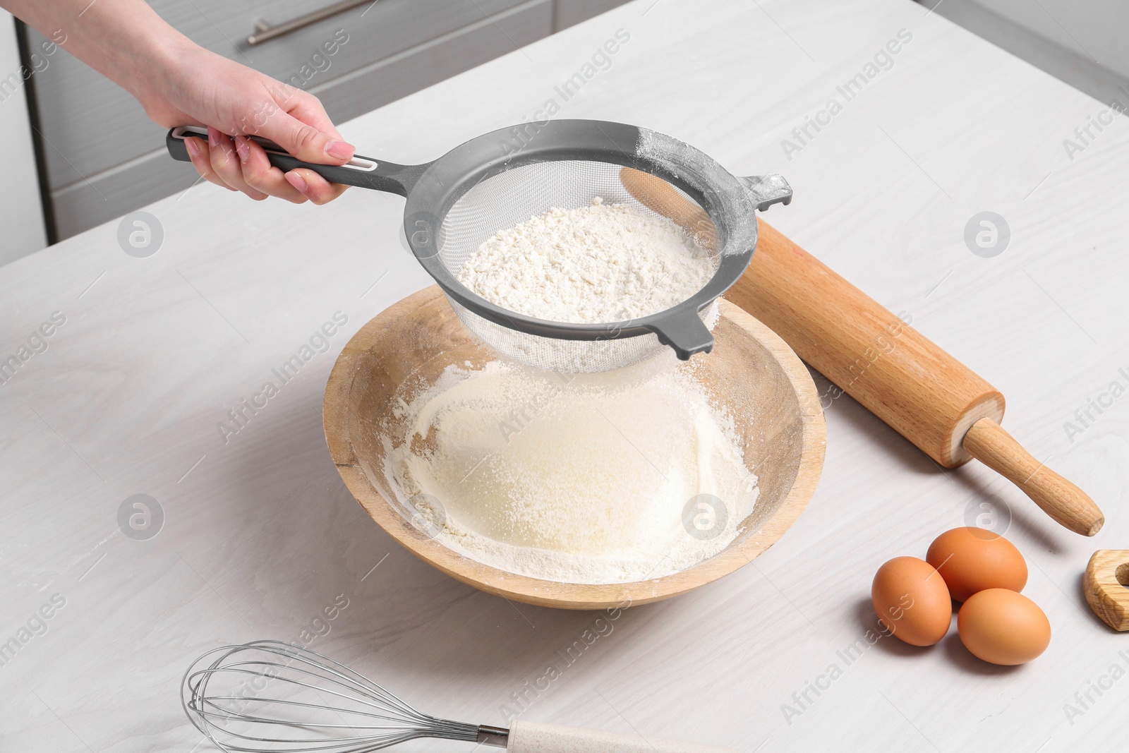 Photo of Woman sieving flour into bowl at white wooden table in kitchen, closeup