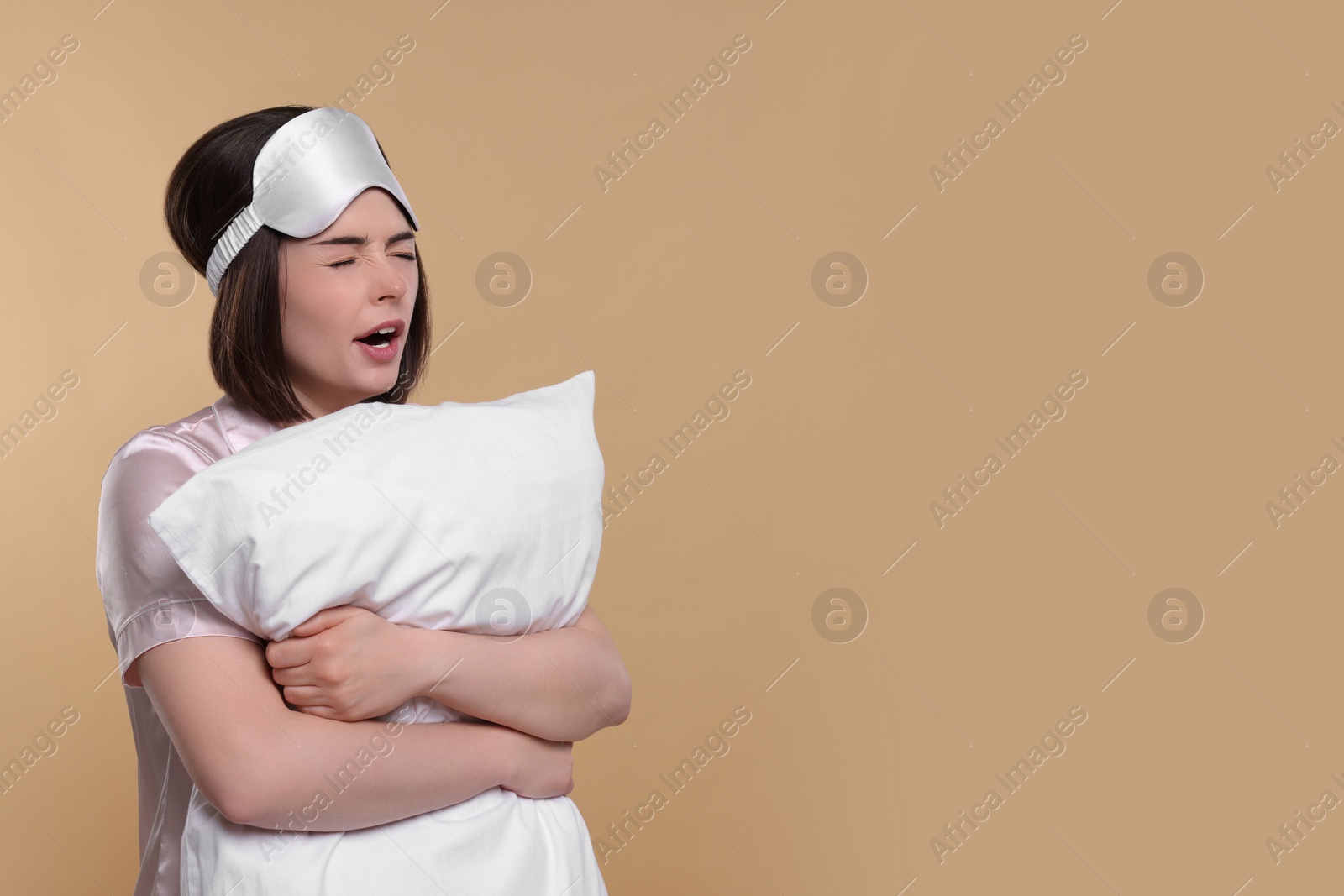 Photo of Tired young woman with sleep mask and pillow yawning on beige background, space for text. Insomnia problem