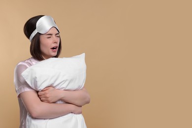 Tired young woman with sleep mask and pillow yawning on beige background, space for text. Insomnia problem