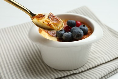 Eating delicious creme brulee with berries from spoon at white table, closeup