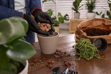 Woman in gloves filling flowerpot with drainage at wooden table indoors, closeup. Transplanting houseplants