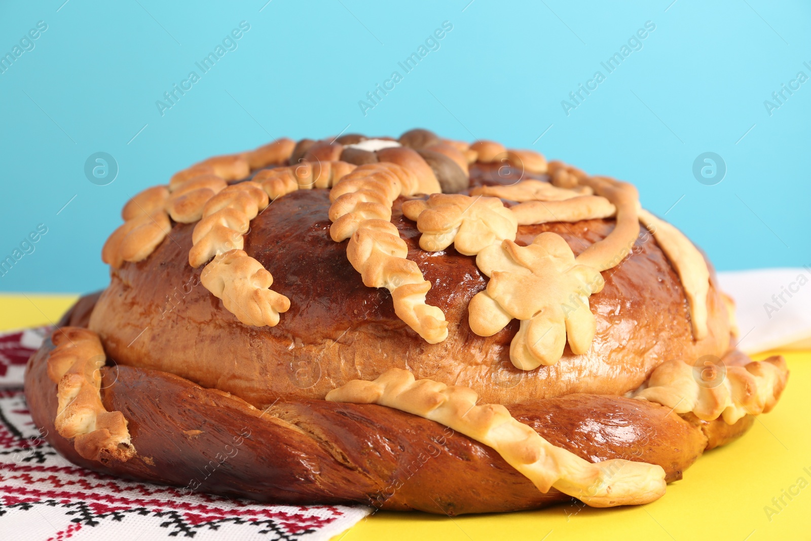 Photo of Rushnyk with korovai on yellow table against light blue background, closeup. Ukrainian bread and salt welcoming tradition