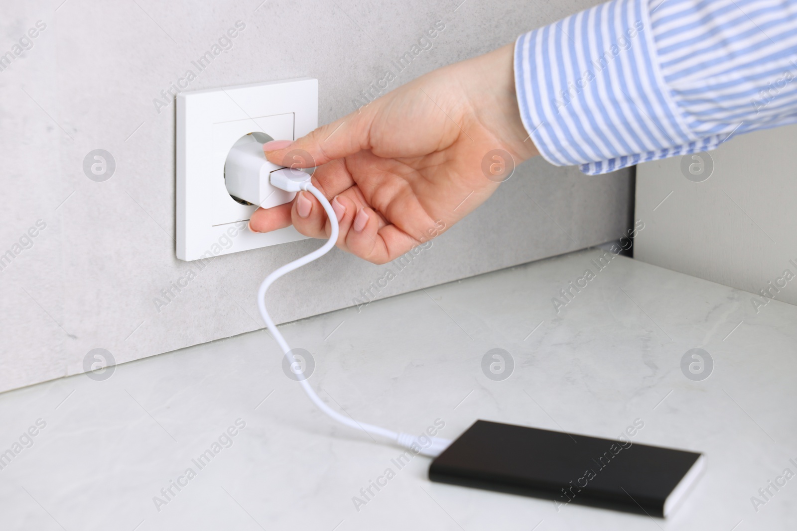 Photo of Woman plugging power bank into socket at white table indoors, closeup