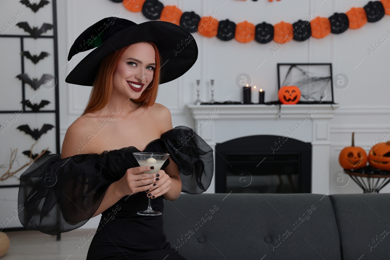 Photo of Happy young woman in scary witch costume with glass of cocktail indoors, space for text. Halloween celebration