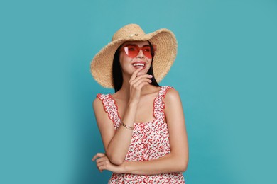 Beautiful young woman with straw hat and heart shaped sunglasses on light blue background