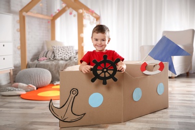 Photo of Cute little boy playing with cardboard boat in bedroom