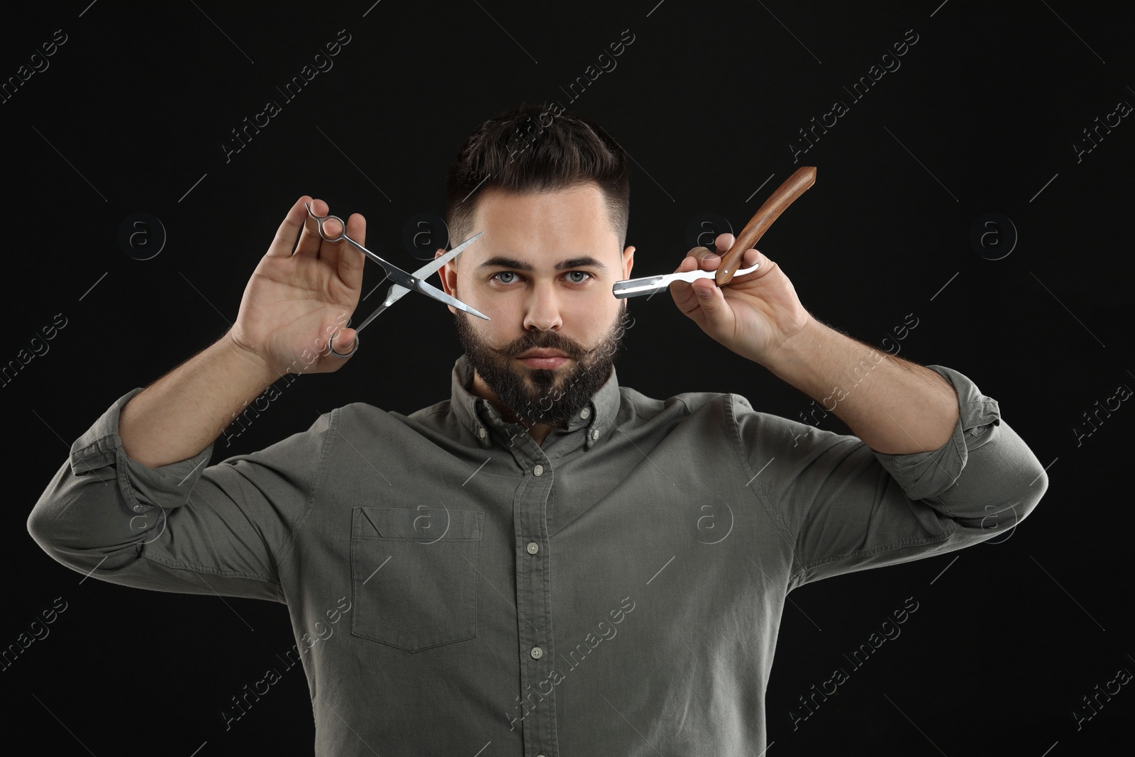 Photo of Handsome young man with mustache holding blade and scissors on black background