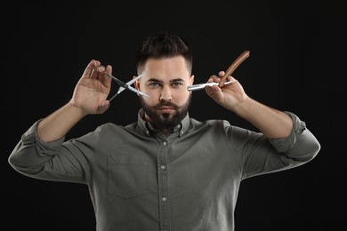 Handsome young man with mustache holding blade and scissors on black background