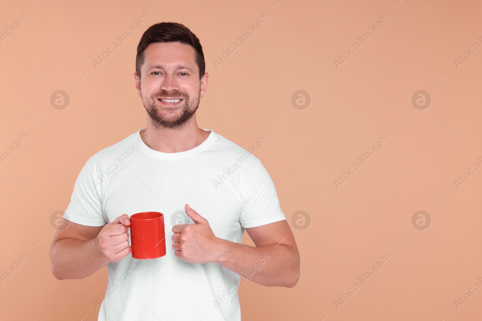 Photo of Portrait of happy man holding red mug and showing thumb up on beige background. Space for text
