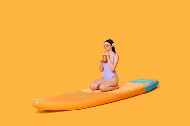 Photo of Happy woman with refreshing drink resting on SUP board against orange background