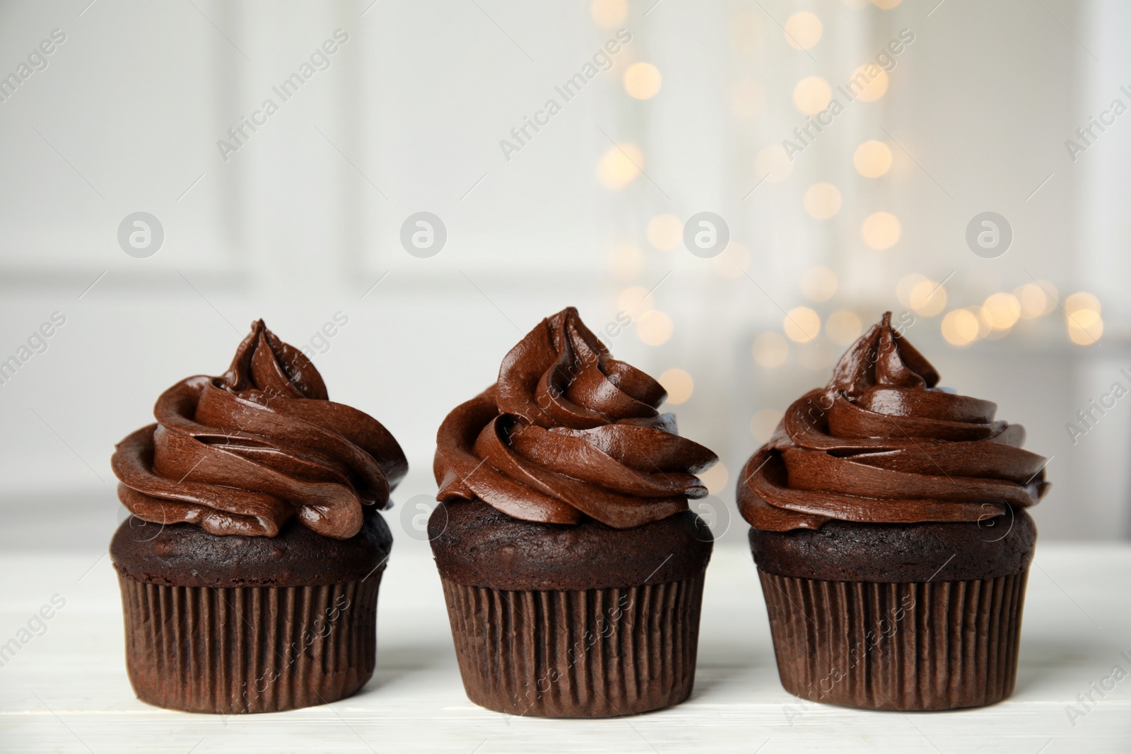 Photo of Delicious chocolate cupcakes with cream on white wooden table against blurred lights. Space for text