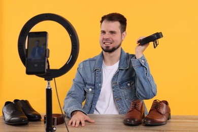 Smiling fashion blogger showing men's accessories while recording video at table against orange background