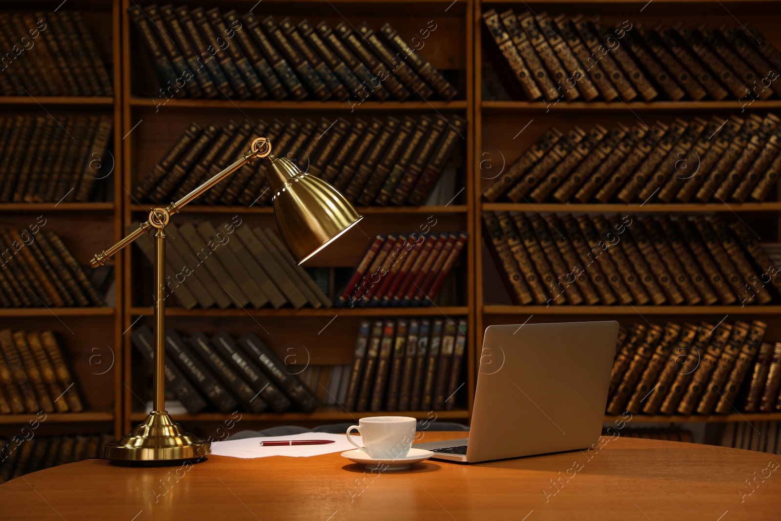 Photo of Lamp, cup of drink and laptop on wooden table near shelves with collection of vintage books in library