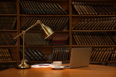 Lamp, cup of drink and laptop on wooden table near shelves with collection of vintage books in library