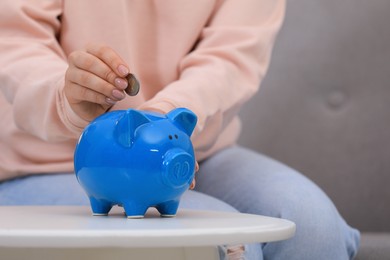 Photo of Young woman putting coin into piggy bank at table indoors, closeup