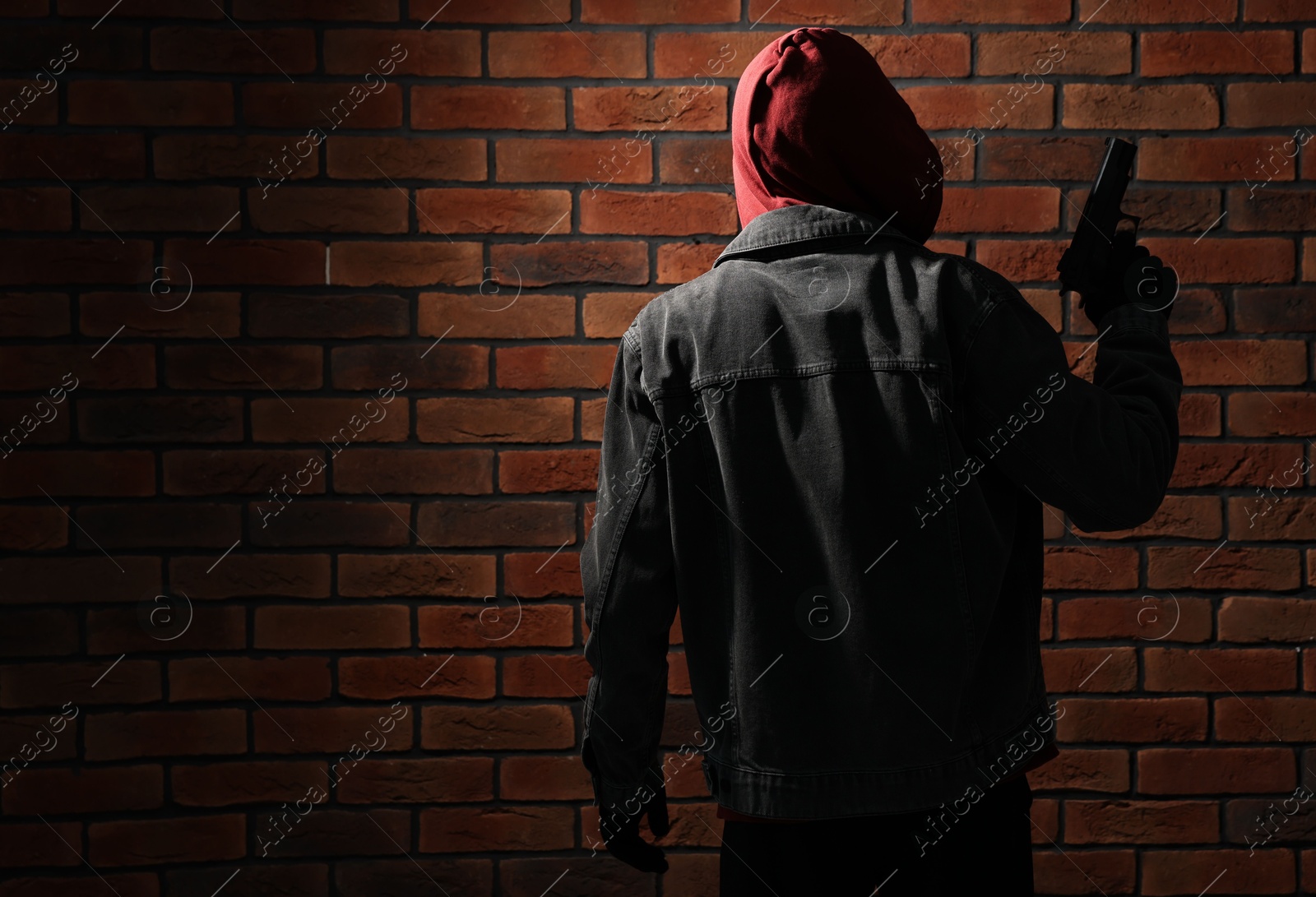 Photo of Thief in hoodie with gun against red brick wall, back view. Space for text