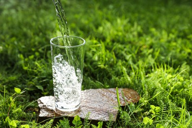 Photo of Pouring fresh water into glass on stone in green grass outdoors. Space for text