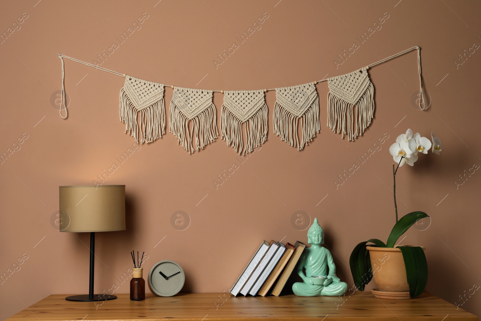 Photo of Potted orchid, decor elements and books on wooden table near brown wall with stylish macrame