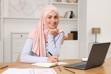 Photo of Muslim woman writing notes near laptop at wooden table in room