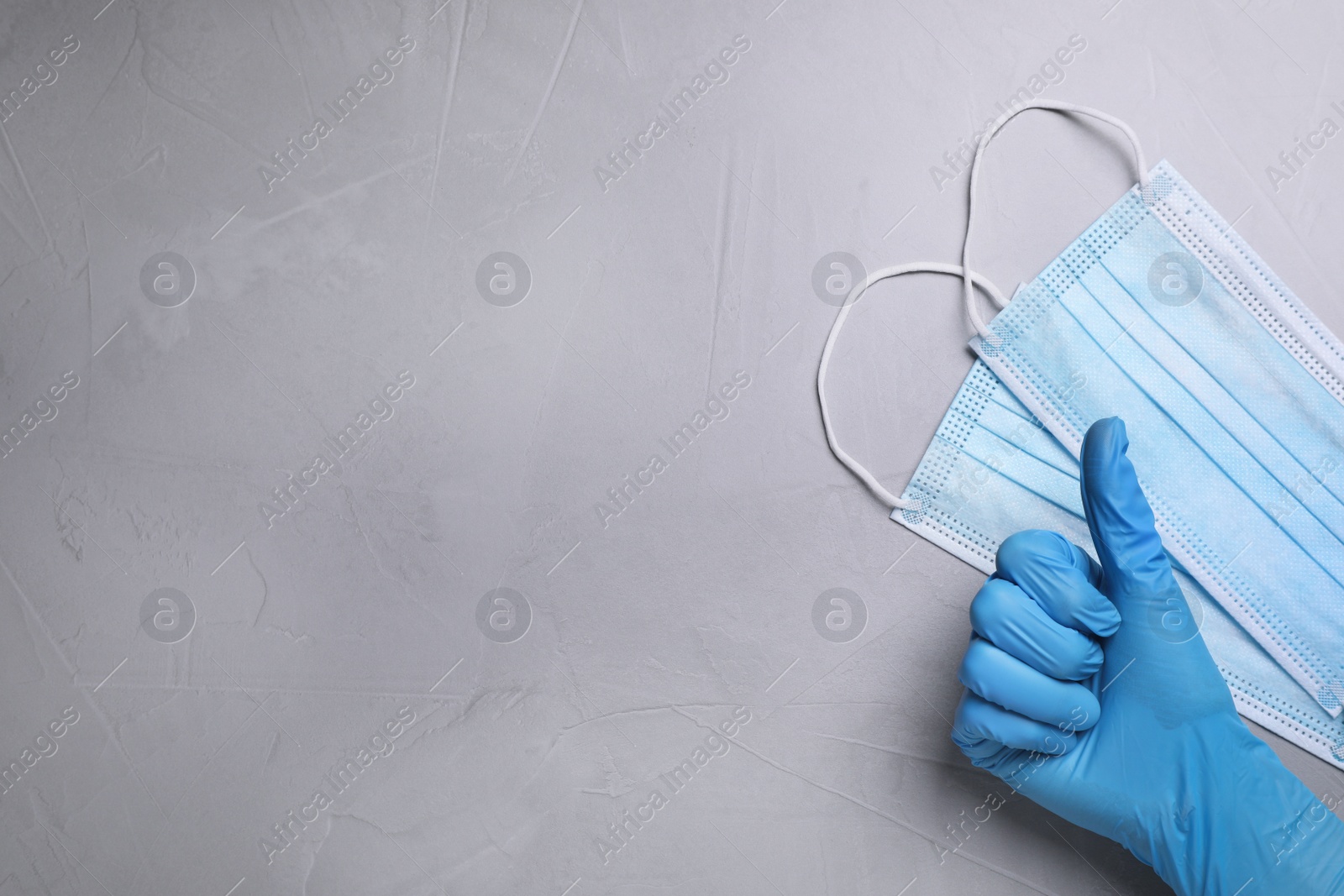 Photo of Doctor showing thumb up near protective masks on light grey stone background, top view with space for text