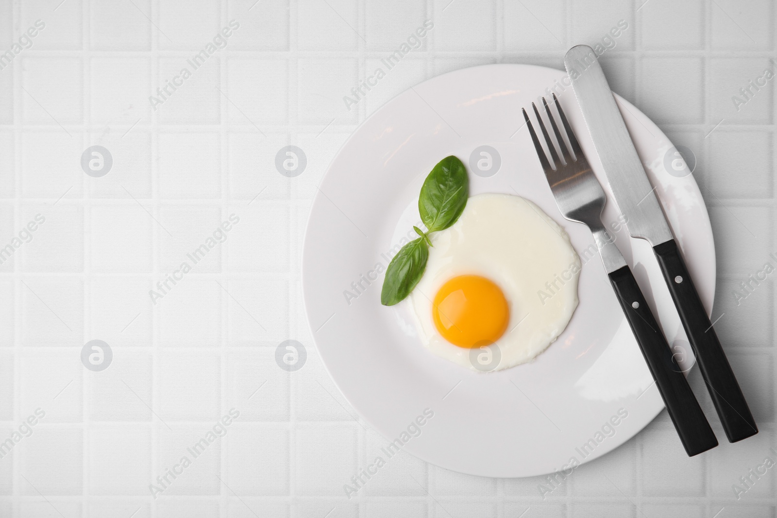 Photo of Tasty fried egg with basil in plate on white tiled table, top view. Space for text