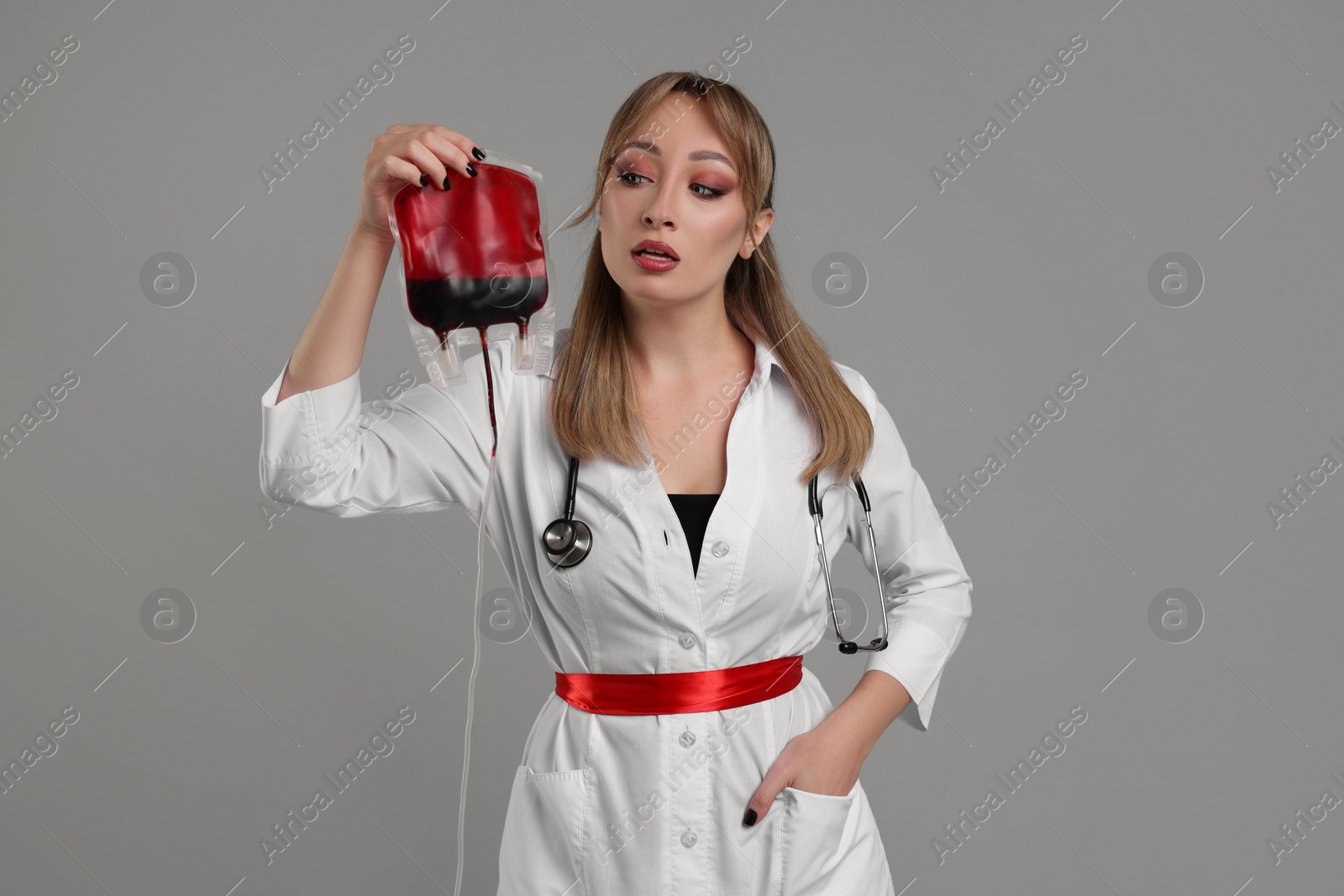 Photo of Woman in scary nurse costume with blood bag on light grey background. Halloween celebration