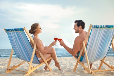 Happy young couple with glasses of wine sitting on deck chairs at sea beach