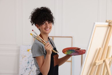 Photo of Young woman holding brushes and artist`s palette near easel with canvas