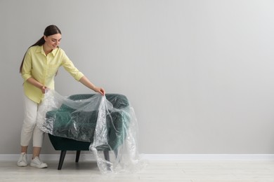 Photo of Young woman putting plastic film away from armchair near light grey wall indoors. Space for text