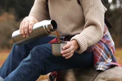Photo of Woman pouring hot drink from metallic thermos into cup lid outdoors, closeup