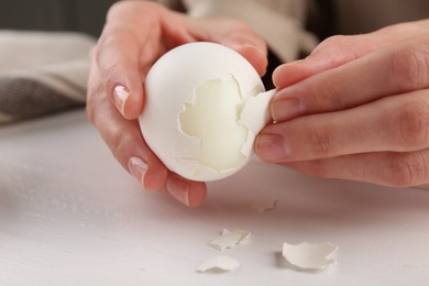 Woman peeling boiled egg at white wooden table, closeup