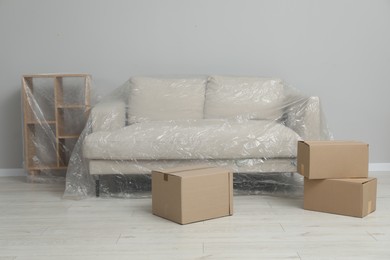 Shelving unit, sofa covered with plastic film and boxes near light grey wall indoors