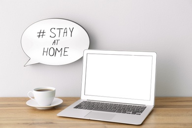 Photo of Laptop, cup of coffee and speech bubble with hashtag STAY AT HOME on white wall. Message to promote self-isolation during COVID‑19 pandemic