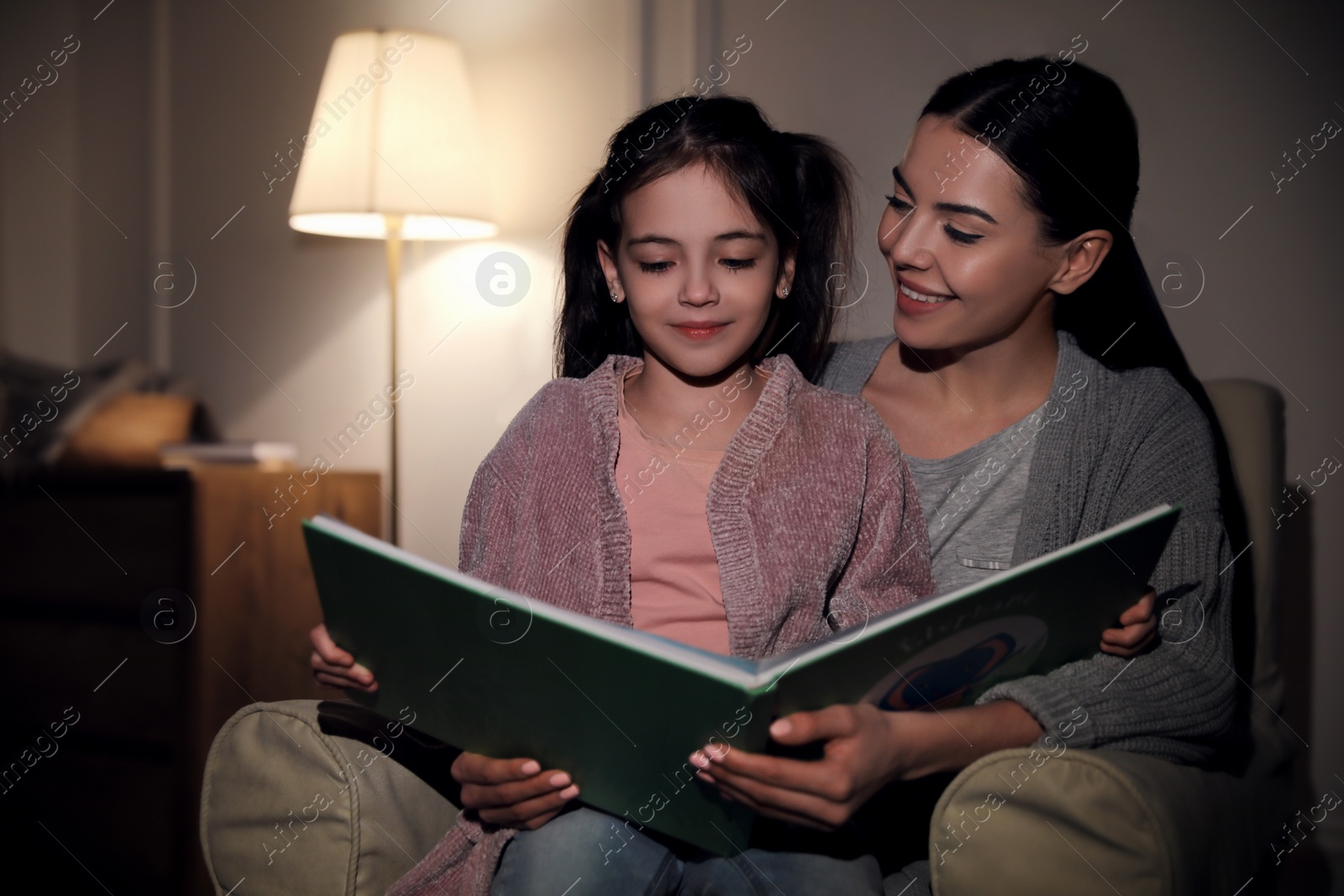 Photo of Little girl with mother reading fairy tale at home in evening