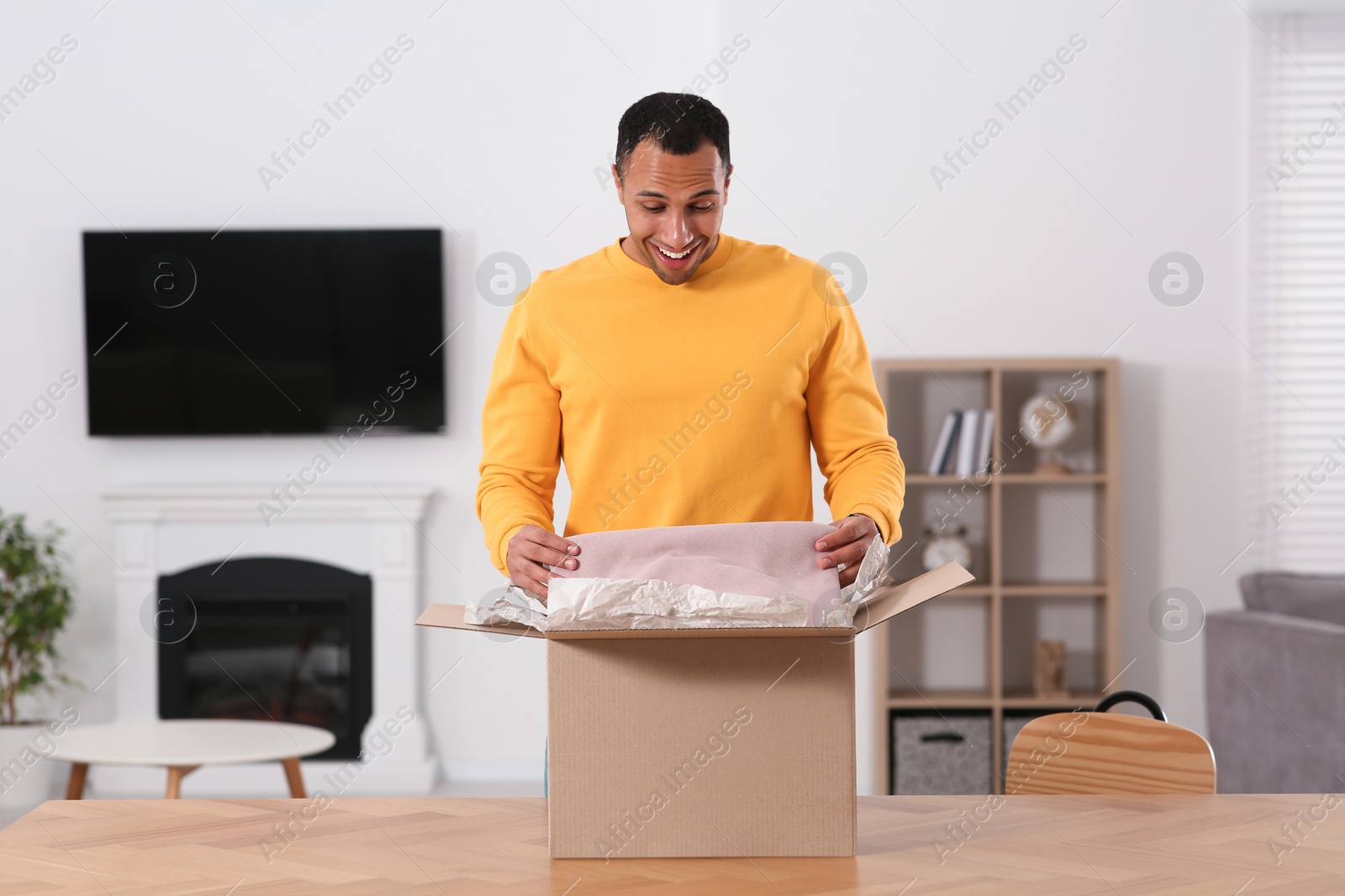 Photo of Happy young man opening parcel at table indoors. Internet shopping