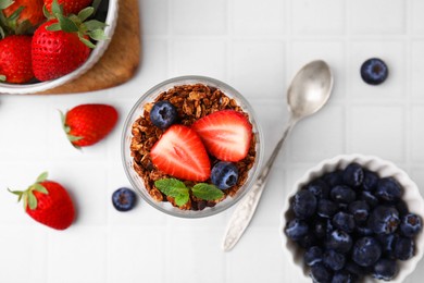 Photo of Tasty granola, strawberries, blueberries and spoon on white tiled table, flat lay