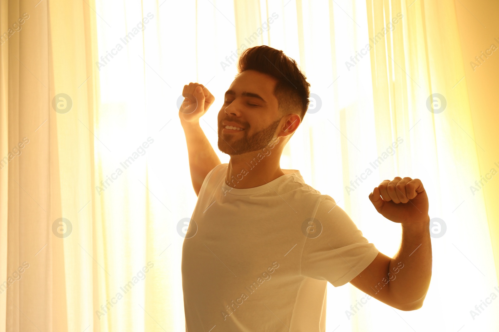 Photo of Young man stretching near window at home. Lazy morning