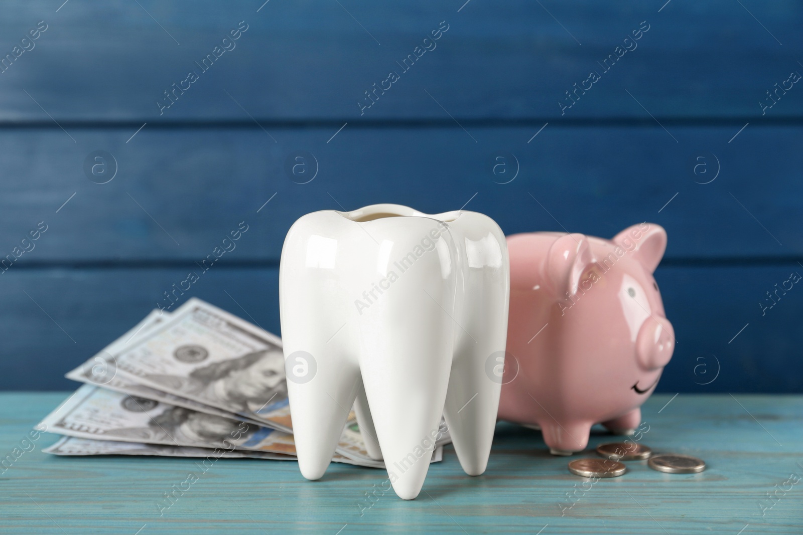 Photo of Ceramic model of tooth, piggy bank and money on light blue wooden table. Expensive treatment