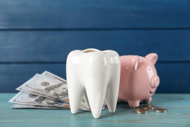 Photo of Ceramic model of tooth, piggy bank and money on light blue wooden table. Expensive treatment