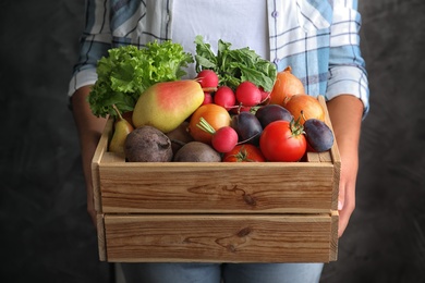 Photo of Farmer holding wooden crate filled with fresh vegetables and fruits on grey background, closeup
