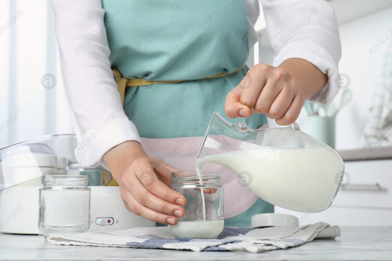 Photo of Woman pouring milk into glass jar at white marble table, closeup. Making yogurt