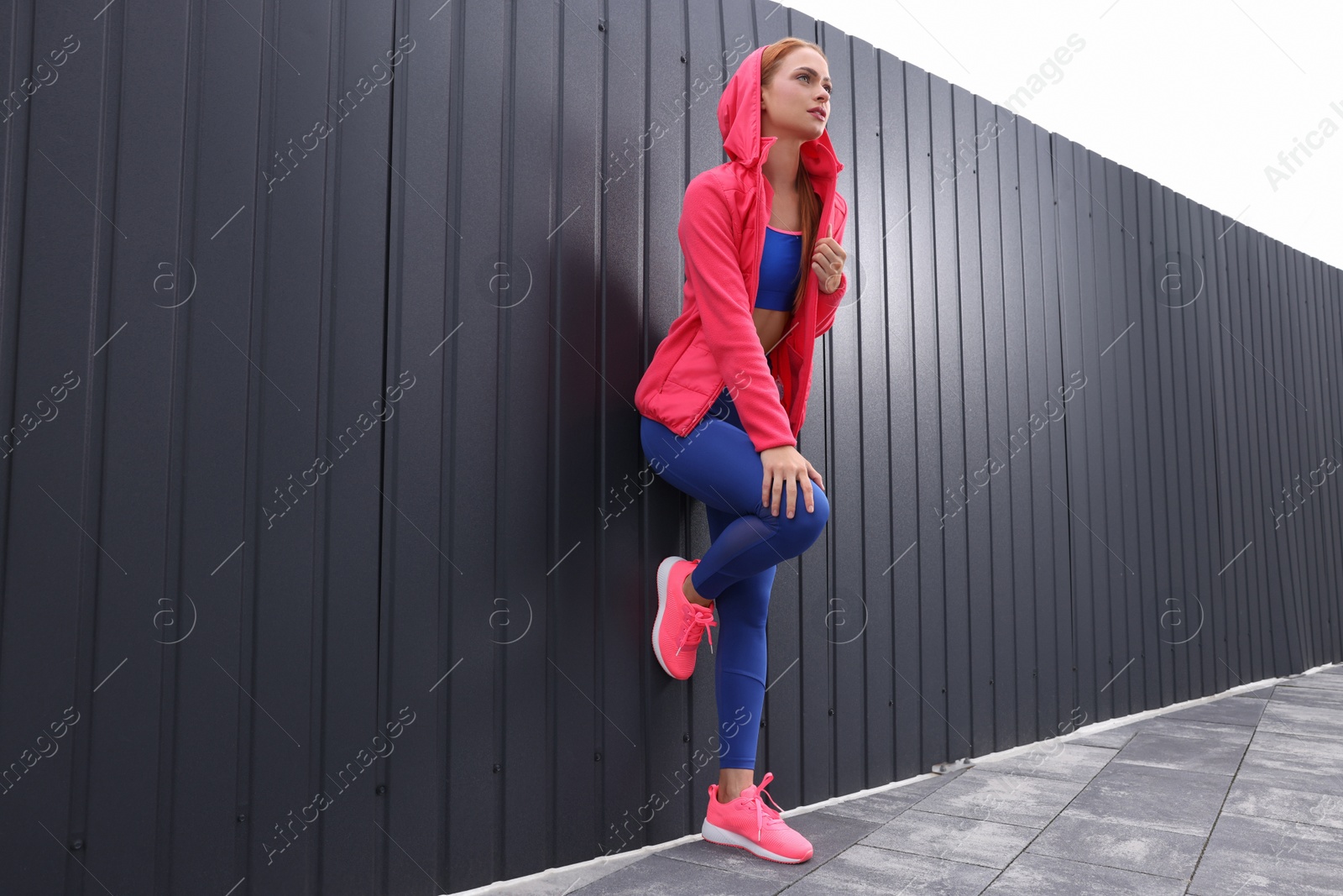 Photo of Beautiful woman in gym clothes posing near dark grey fence on street, low angle view