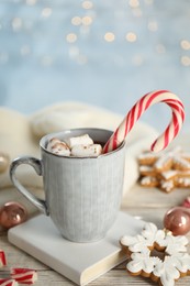 Photo of Cup of tasty cocoa with marshmallows and Christmas candy cane on white wooden table against blurred festive lights