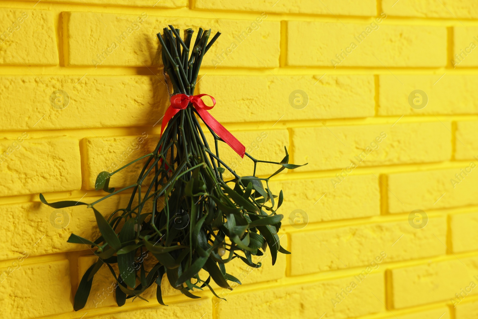 Photo of Mistletoe bunch with red bow hanging on yellow brick wall, space for text. Traditional Christmas decor