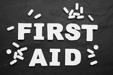 Photo of Words First Aid made of white letters and pills on black table, flat lay