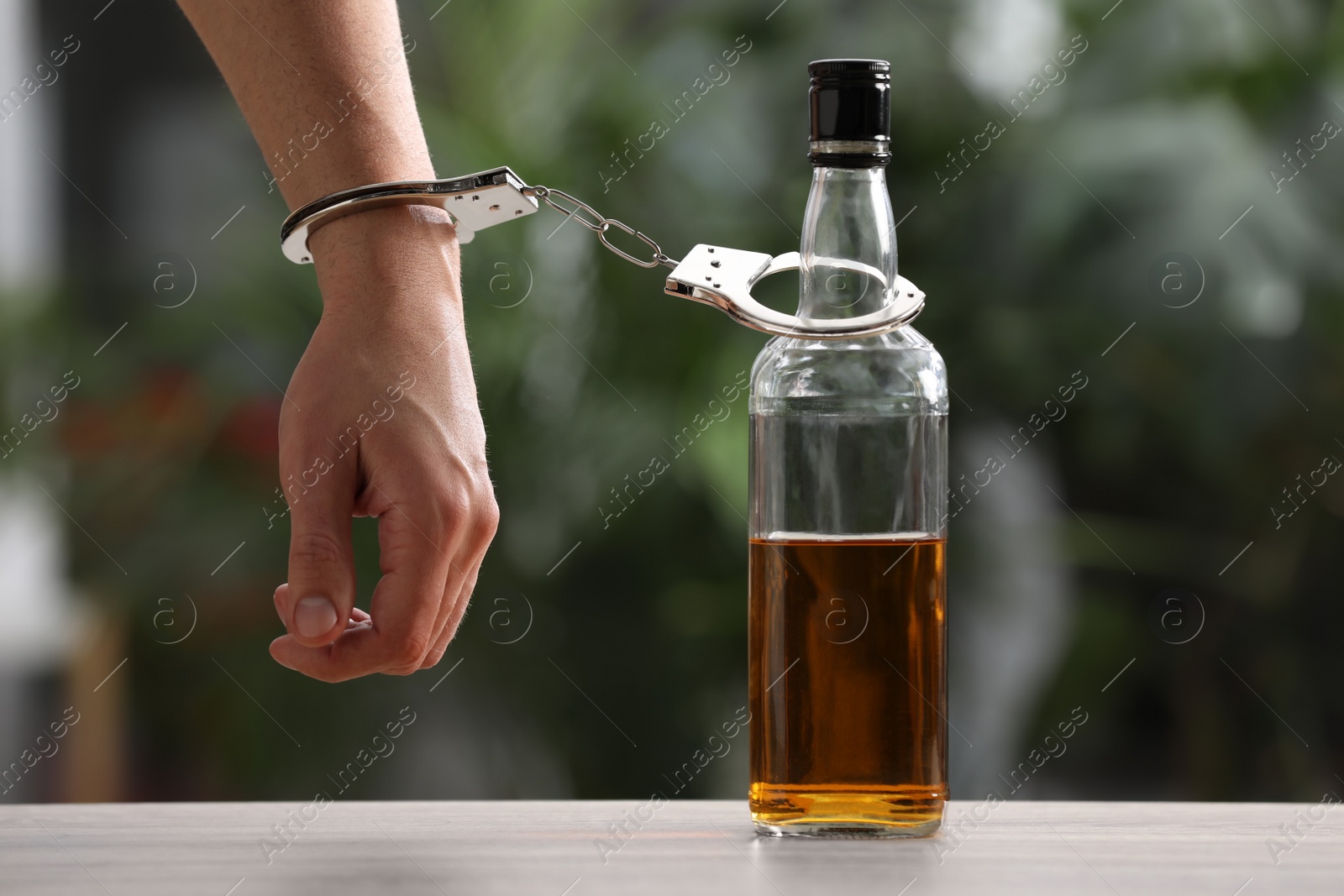 Photo of Addicted man in handcuffs with bottle of alcoholic drink at table against blurred background, closeup