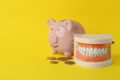 Educational dental typodont model, piggy bank and coins on yellow background, space for text. Expensive treatment